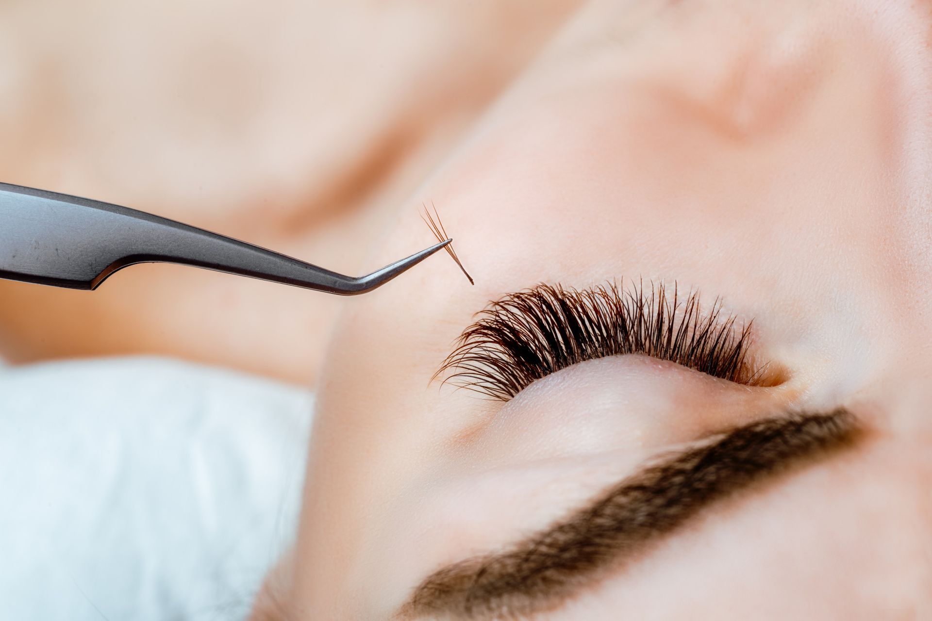 A woman receiving eyelash extensions. Tweezers hold a small cluster of lashes, next to her closed eye.