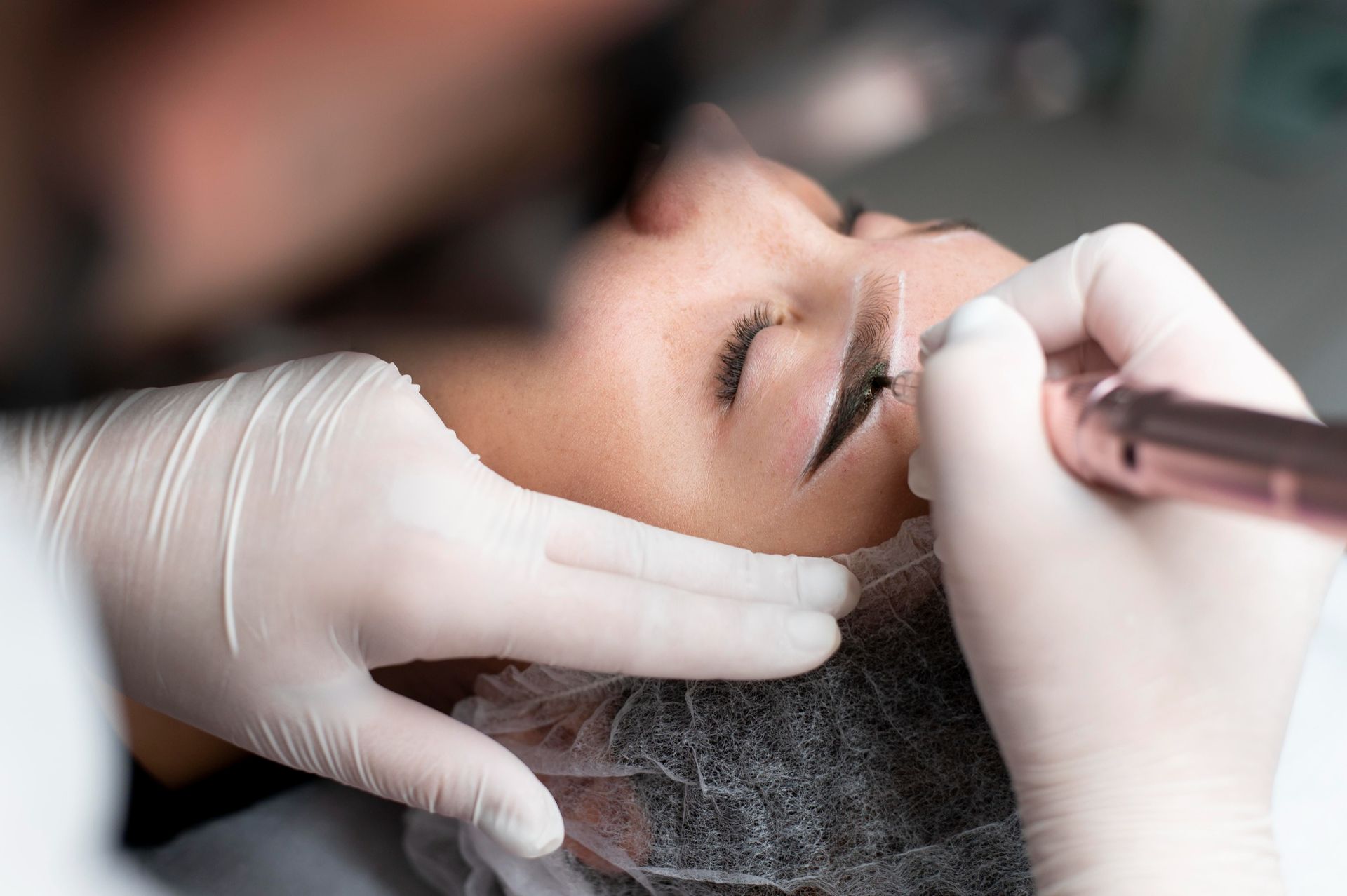 Woman receiving eyebrow microblading. Person's hand in glove holds tool, close-up.