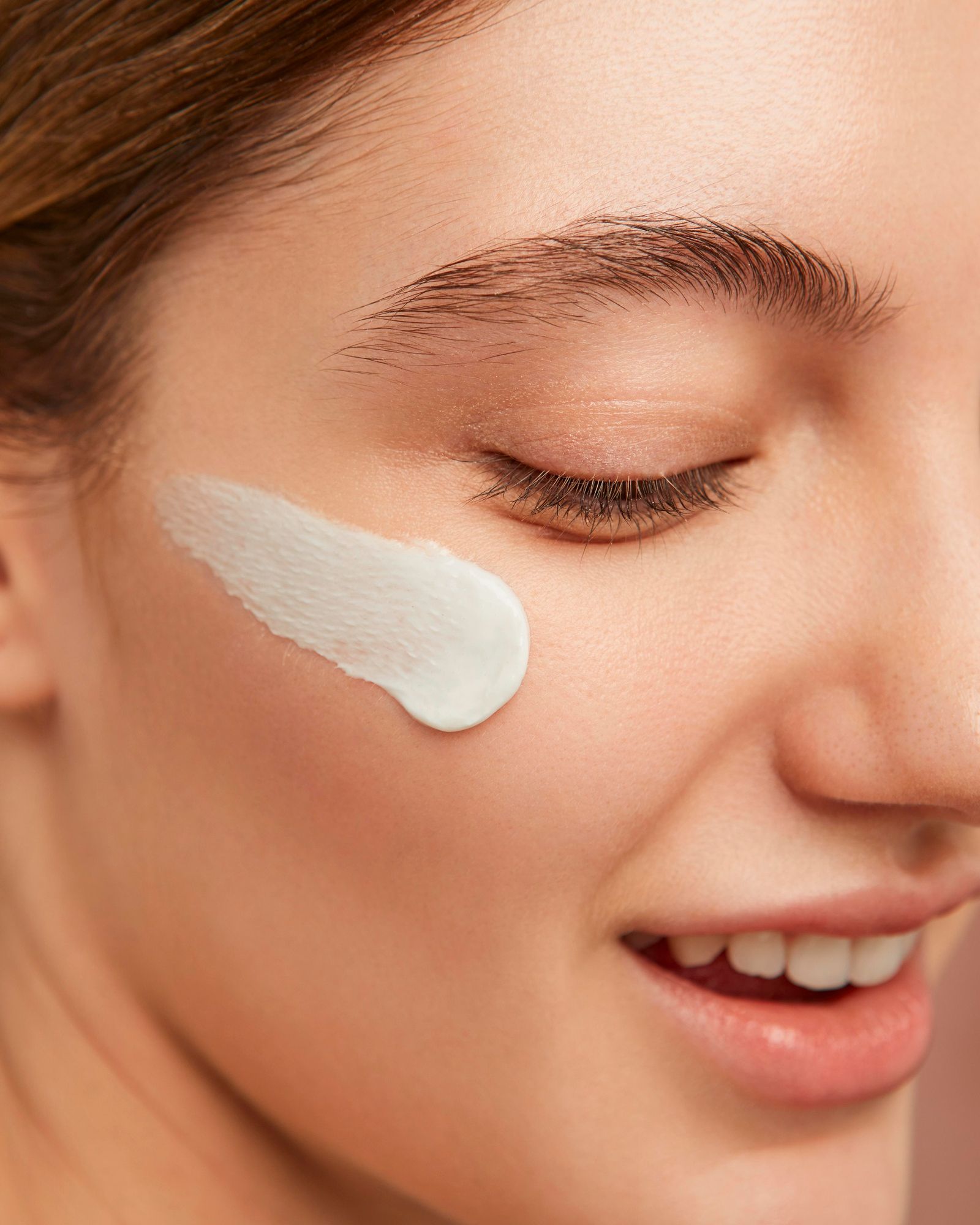 Woman applying cream to her cheek; smiling, closed eyes; light-colored cream.
