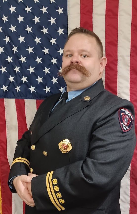 A man with a mustache is standing in front of an American flag.