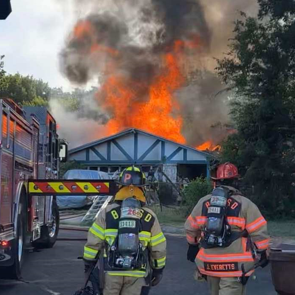 Two firefighters are standing in front of a large house fire.