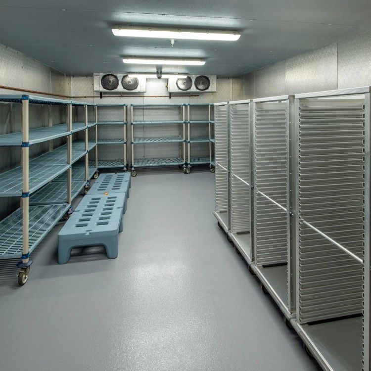 A walk-in commercial refrigerator with grey shelving, empty tray racks, and a blue plastic floor grate.
