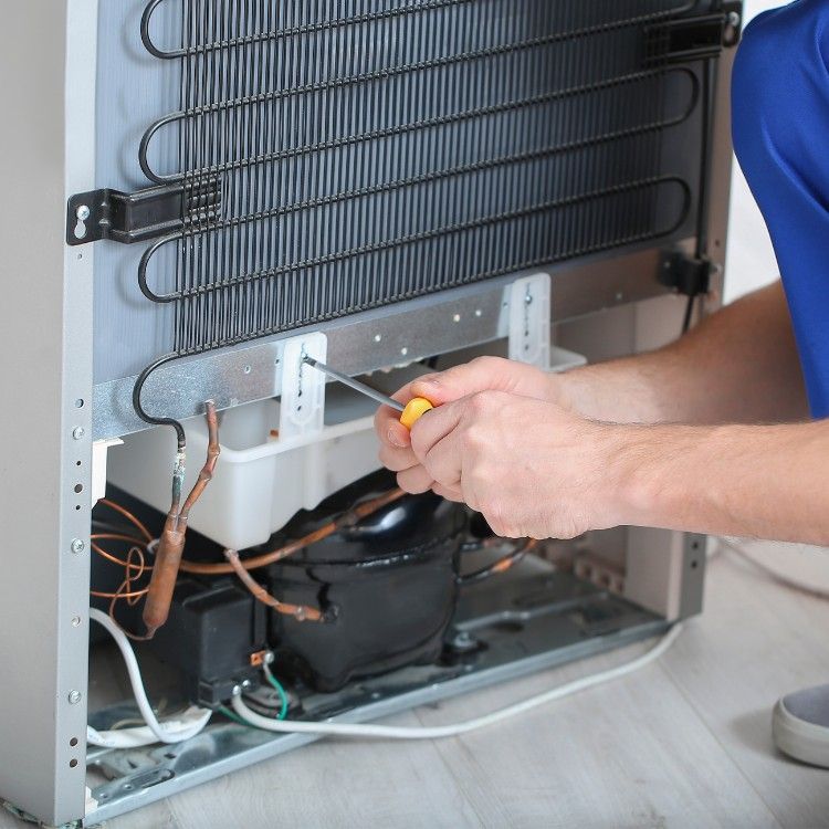 A person in blue work pants uses a screwdriver to repair the back of a refrigerator, exposing the coils and compressor.