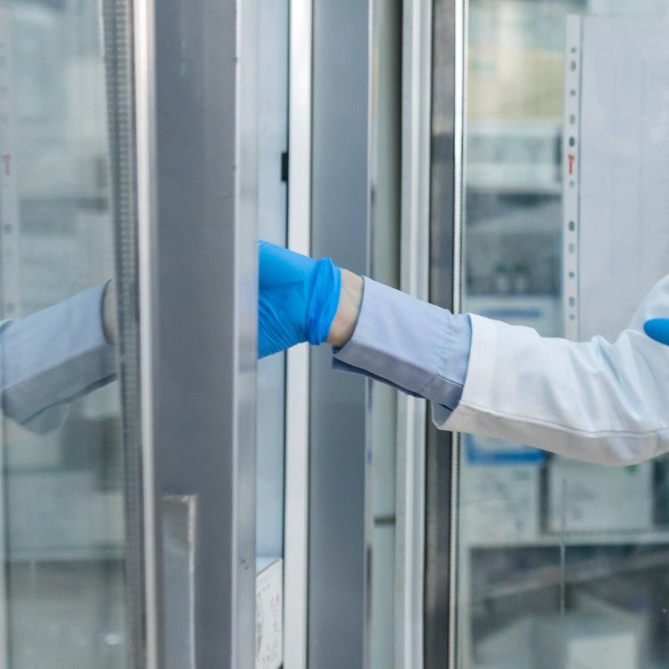 A person in a white lab coat and blue gloves opens a glass-door laboratory refrigerator.