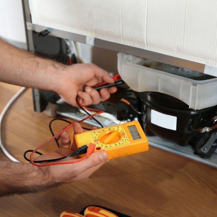 A person using a yellow digital multimeter to test electrical components on the back of a refrigerator.