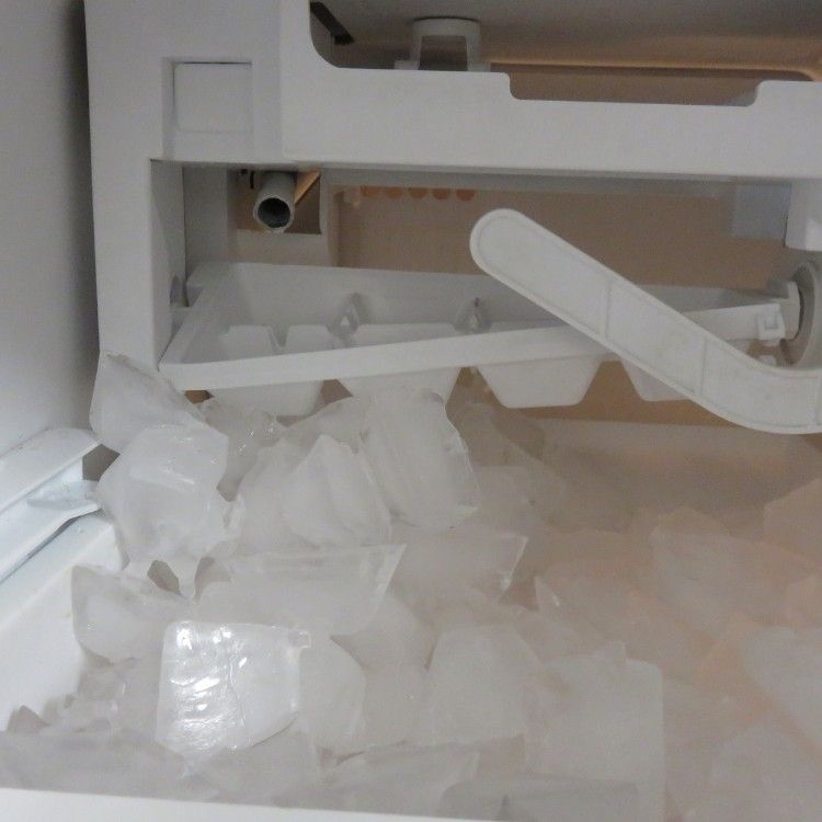 An ice maker inside a freezer, with a white tray filled with ice cubes above a bin overflowing with ice.