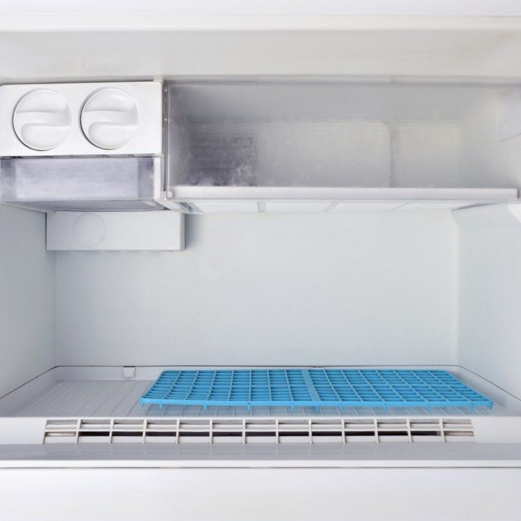 The interior of an empty white freezer with an ice maker on the left and a blue silicone ice cube tray on the floor.