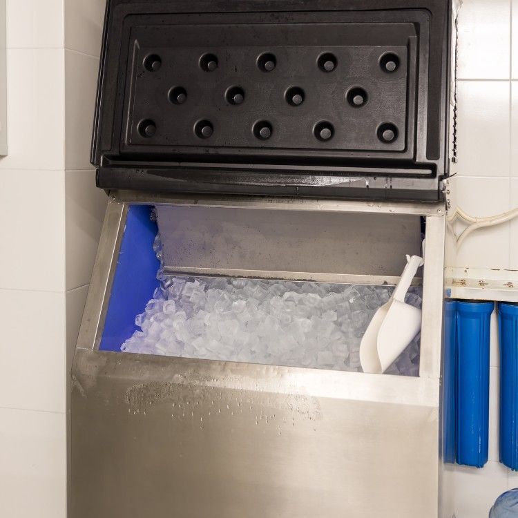 An open stainless steel ice machine filled with ice, featuring a white plastic scoop inside and blue water filters nearby.