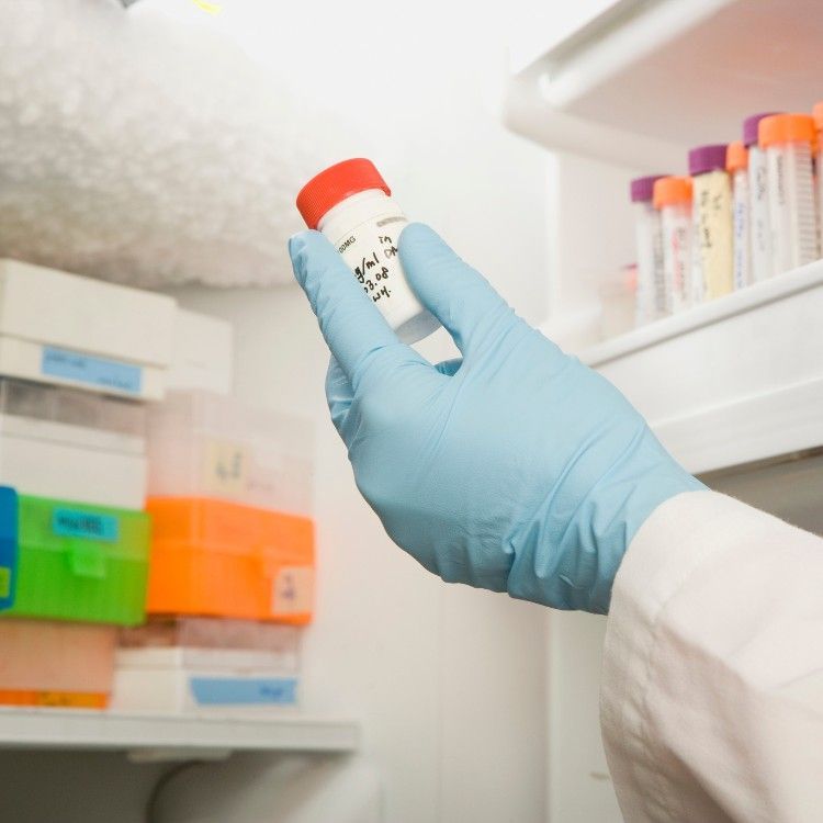 A gloved hand holds a small, labeled sample vial in front of shelves stocked with medical containers in a laboratory.