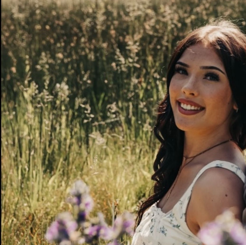 Woman with dark hair smiles, in a field of wildflowers. She wears a white floral top.