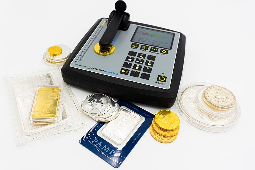 A metal analysis device with assorted gold and silver bars and coins on a white surface.