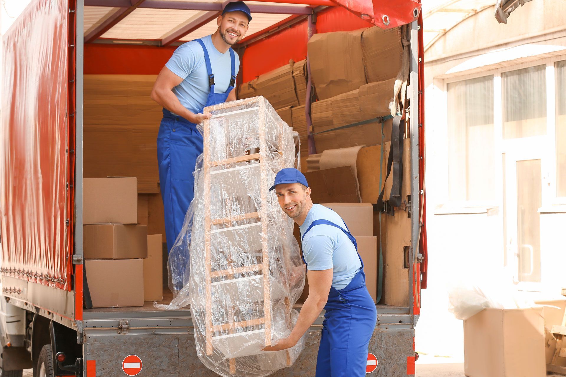 Two movers unloading a large wrapped mirror from a red truck.