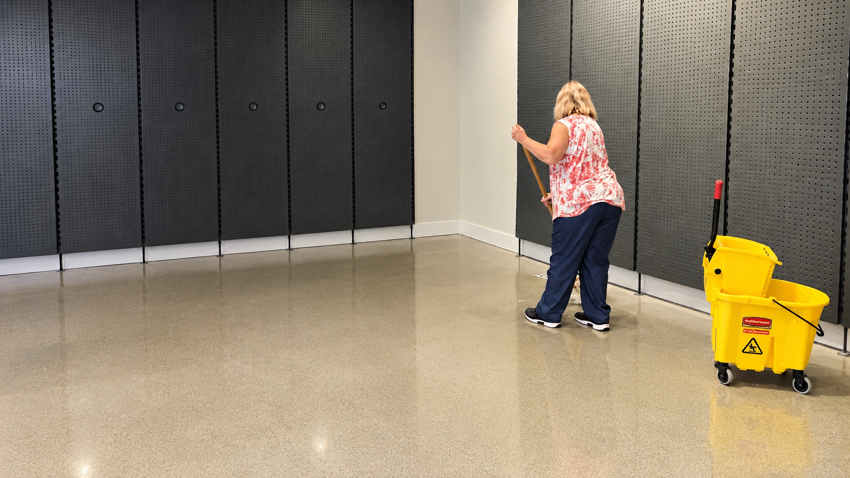 Person in a pink shirt cleaning a hallway floor beside a yellow mop bucket and gray wall panels