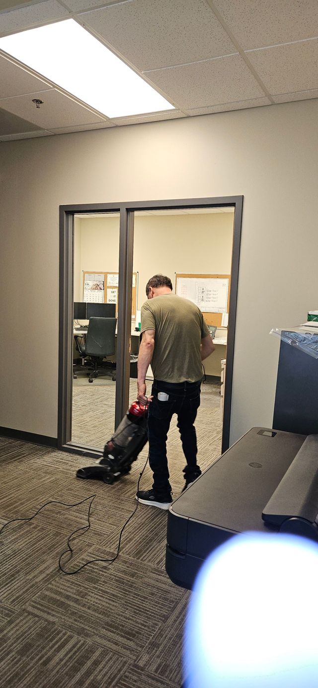Person in an office doorway vacuuming carpet near a desk and blue storage cabinet.