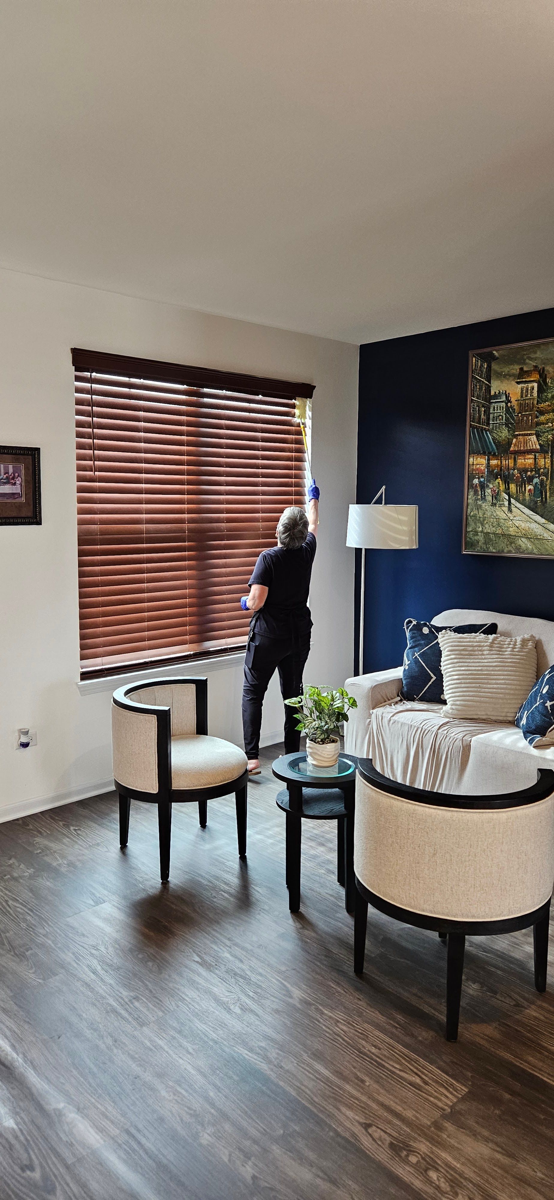 Person stands by a window with blinds in a modern living room, holding a phone.