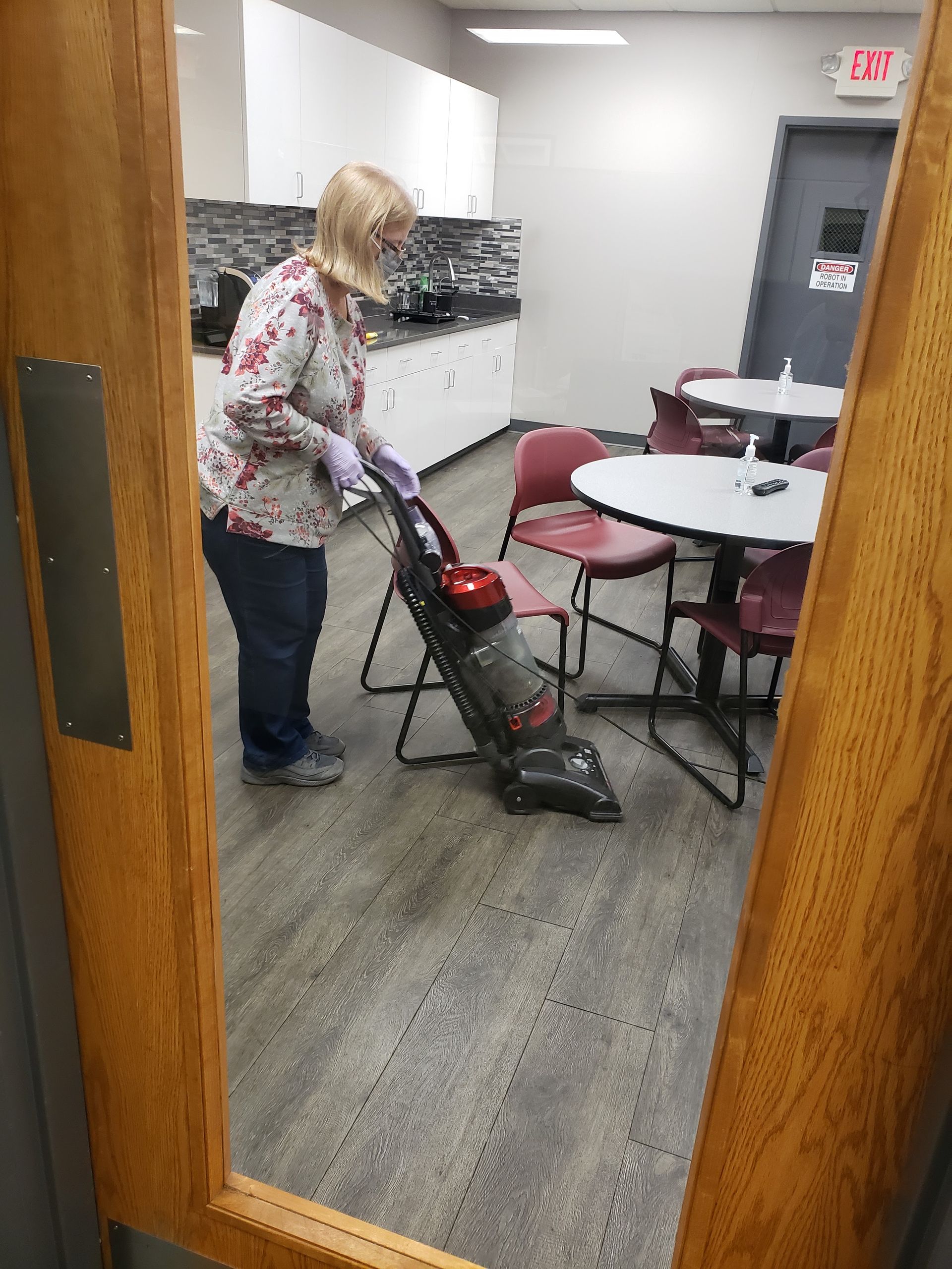 Person vacuuming a carpeted office hallway near round tables and mailboxes.