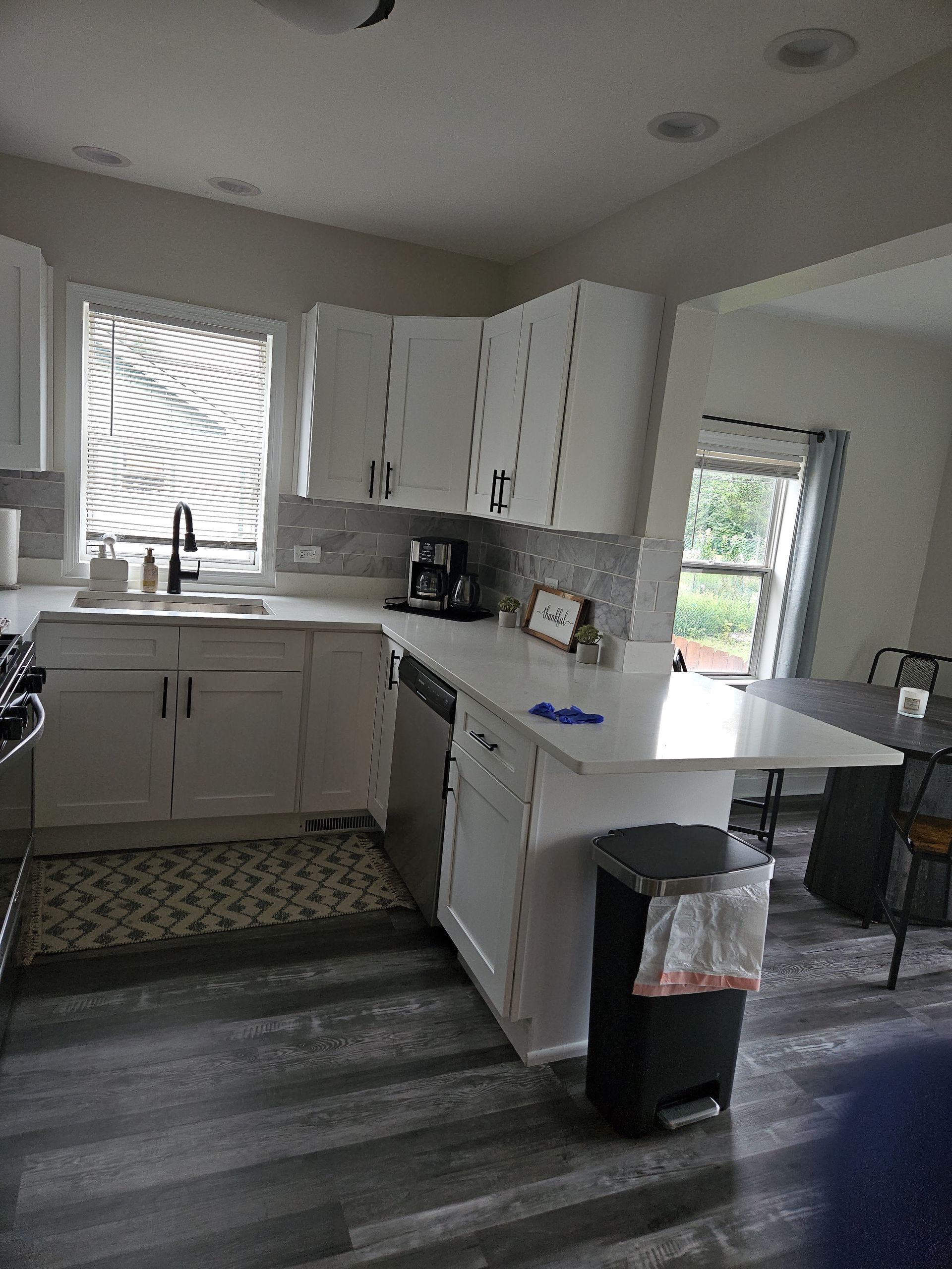 Bright kitchen with white cabinets, sink, island, patterned rug, and dark wood floors; dining area in back.
