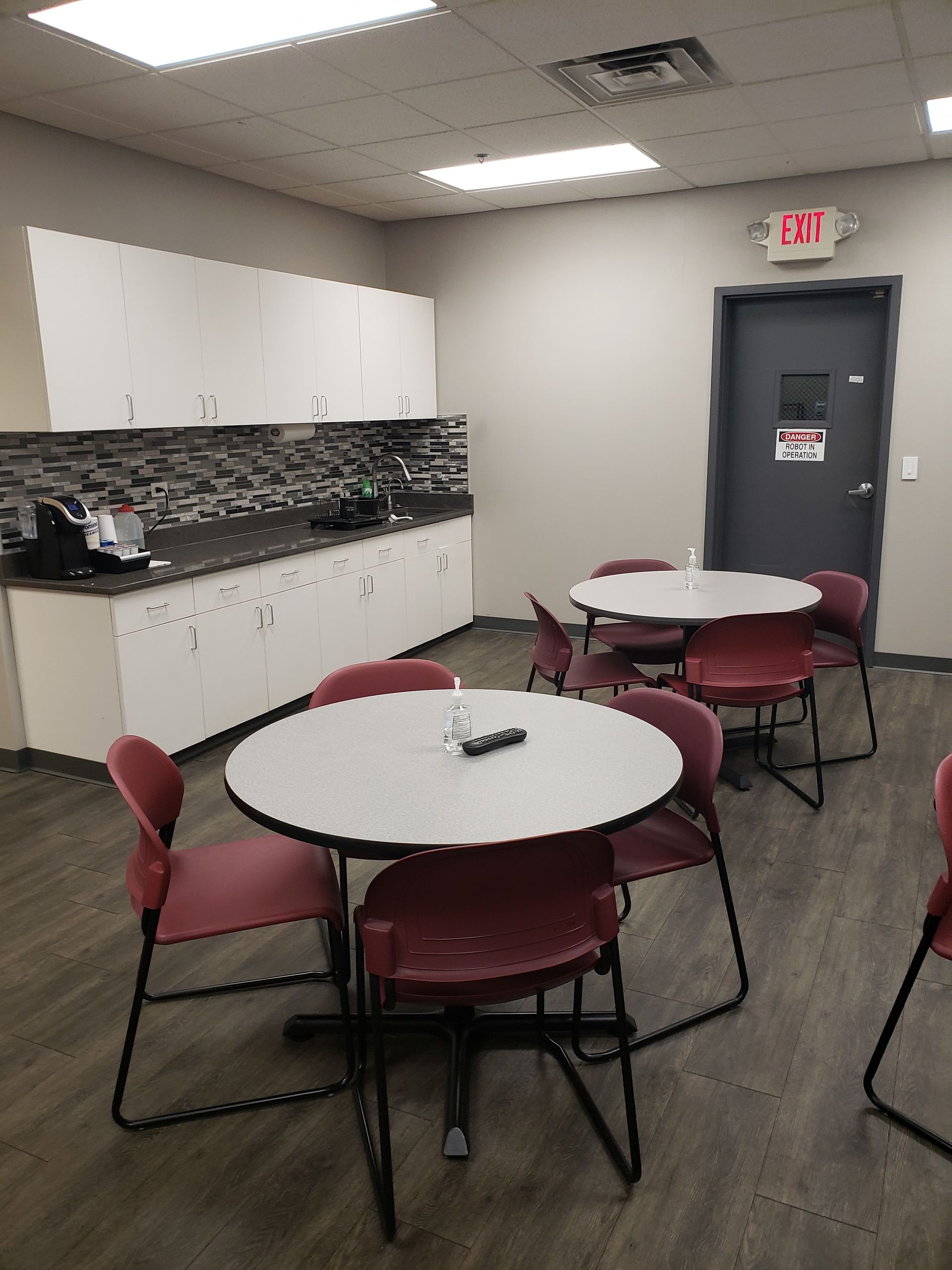 Break room with round tables, red chairs, cabinets, and a dark door under fluorescent lights.