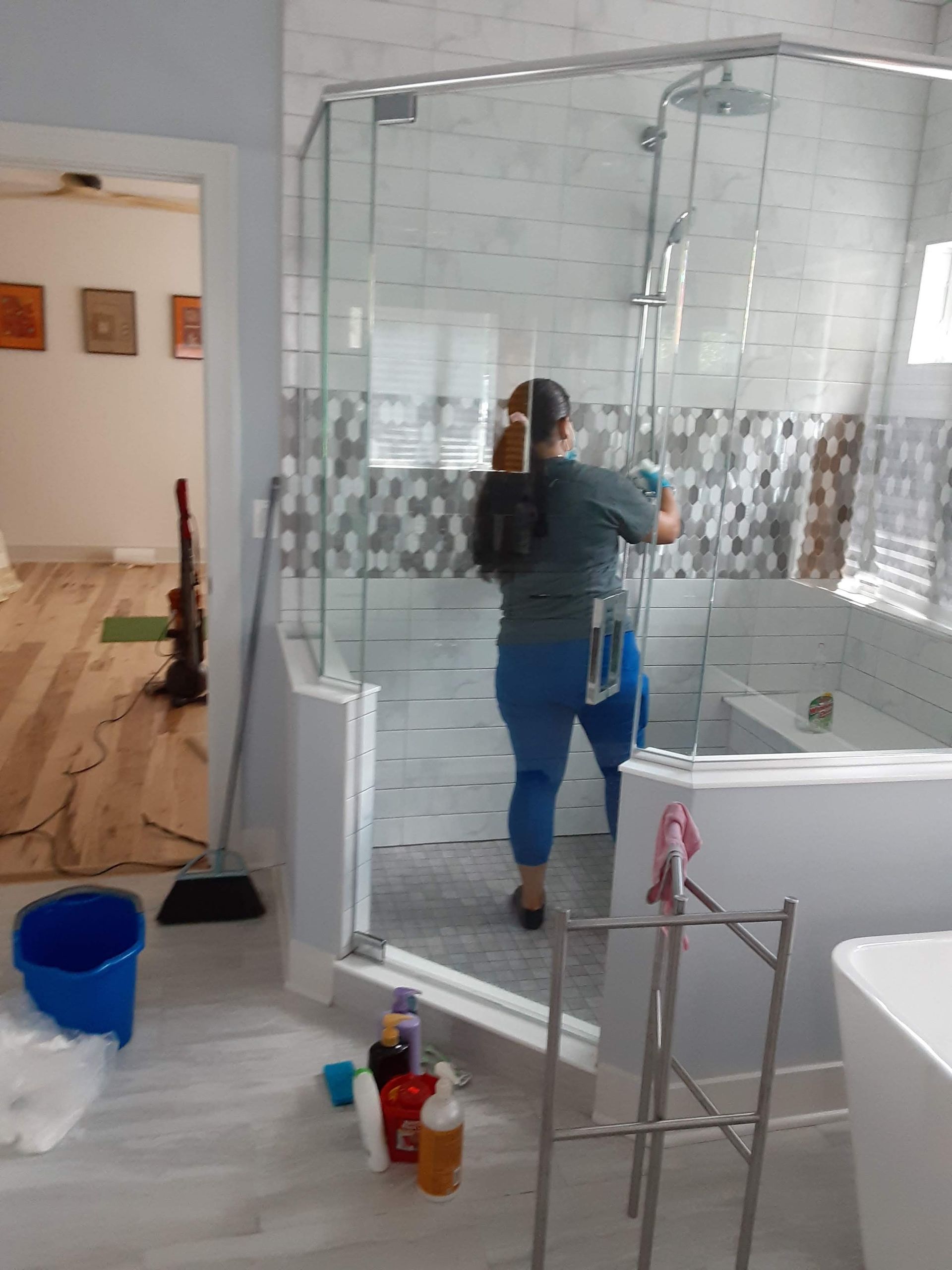 Person cleaning a glass shower enclosure in a tiled bathroom with cleaning supplies nearby