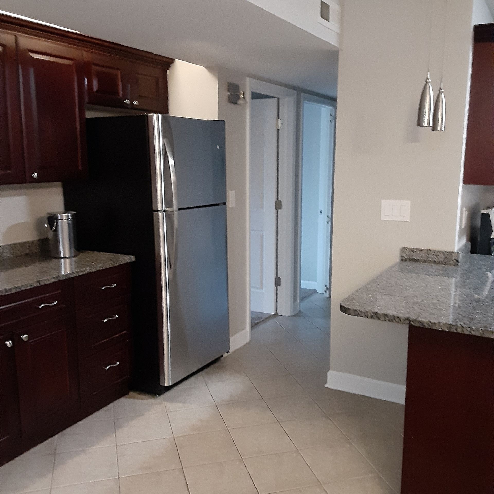 Kitchen with dark wood cabinets, stainless steel fridge, and granite countertops leading to a hallway.