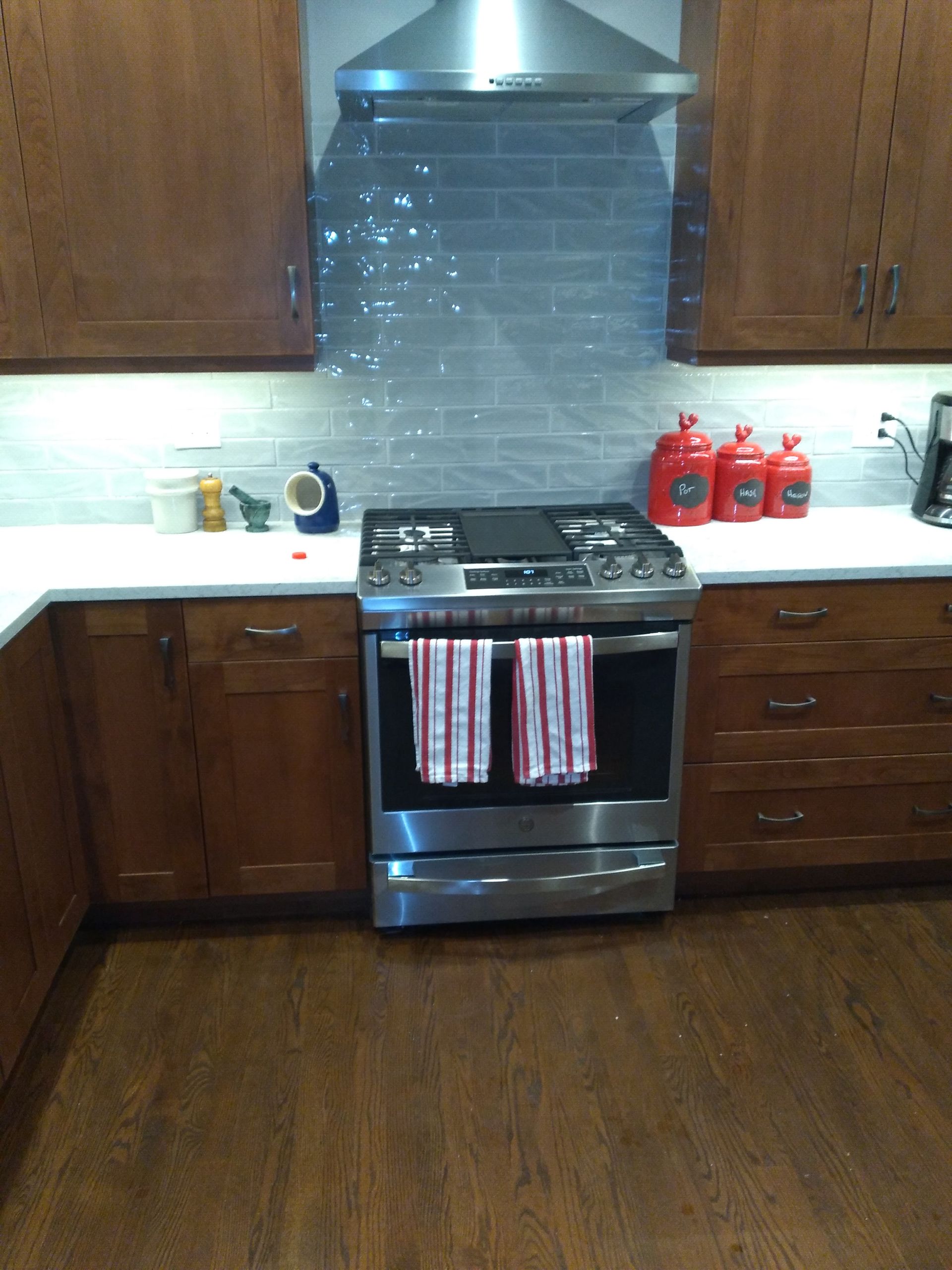 Modern kitchen with a stainless steel stove, wood cabinets, white countertops, and red bottles on the right.