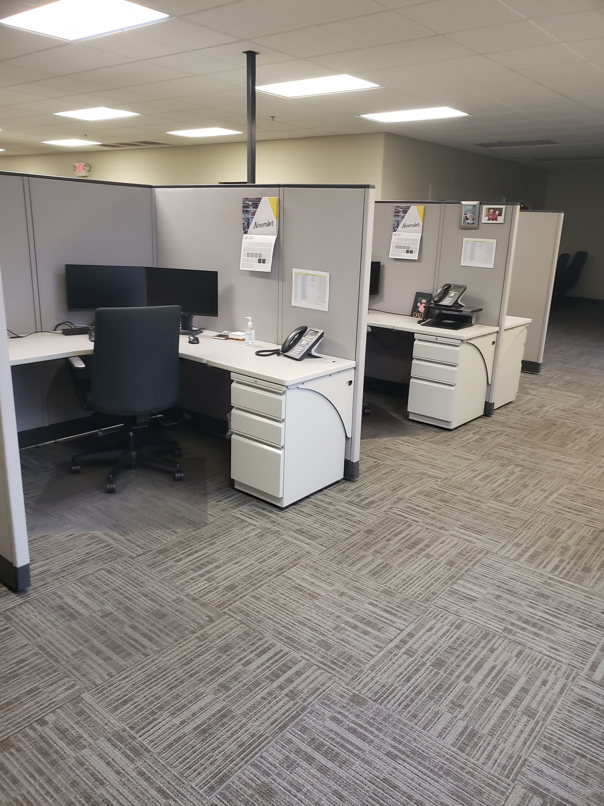 Empty office cubicles with desks, black chairs, and overhead fluorescent lights in a carpeted workspace.