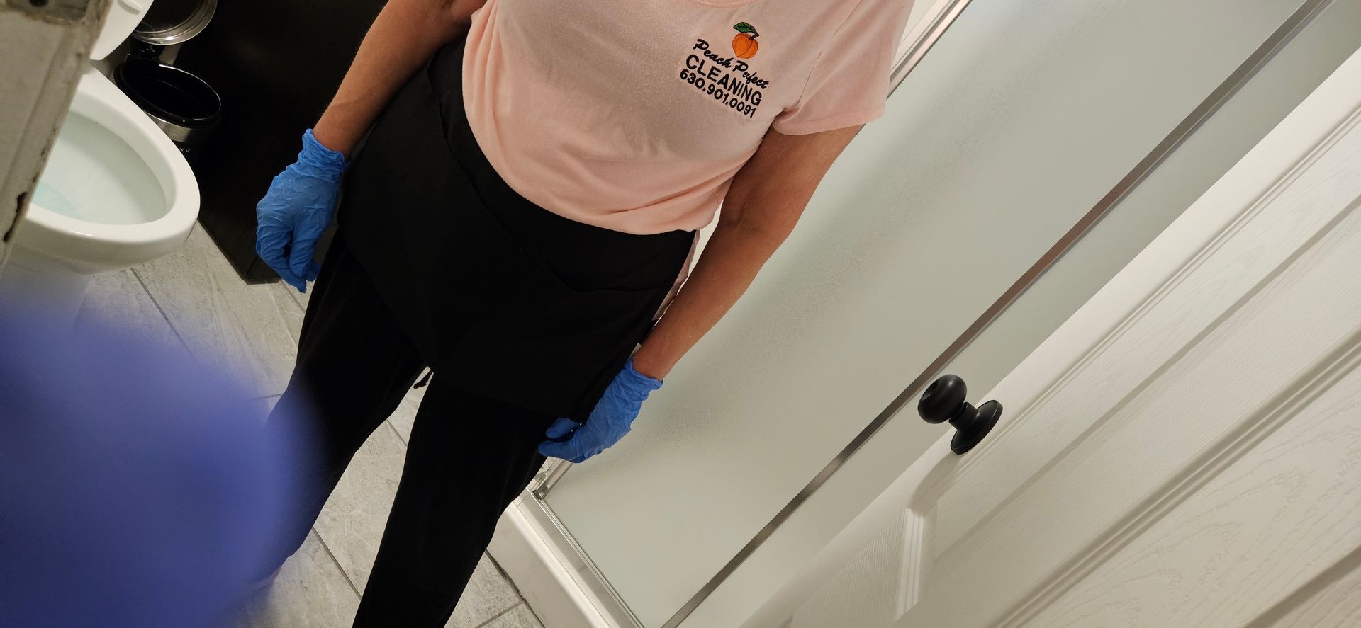 Person in pink shirt and blue gloves standing in a bathroom near a sink and white doors.