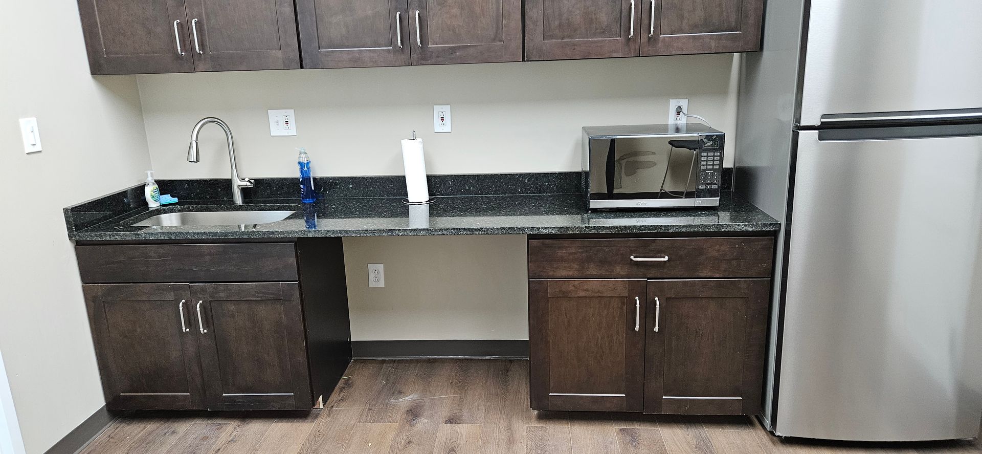 Small kitchen with dark wood cabinets, black countertop, sink, microwave, and stainless steel refrigerator.