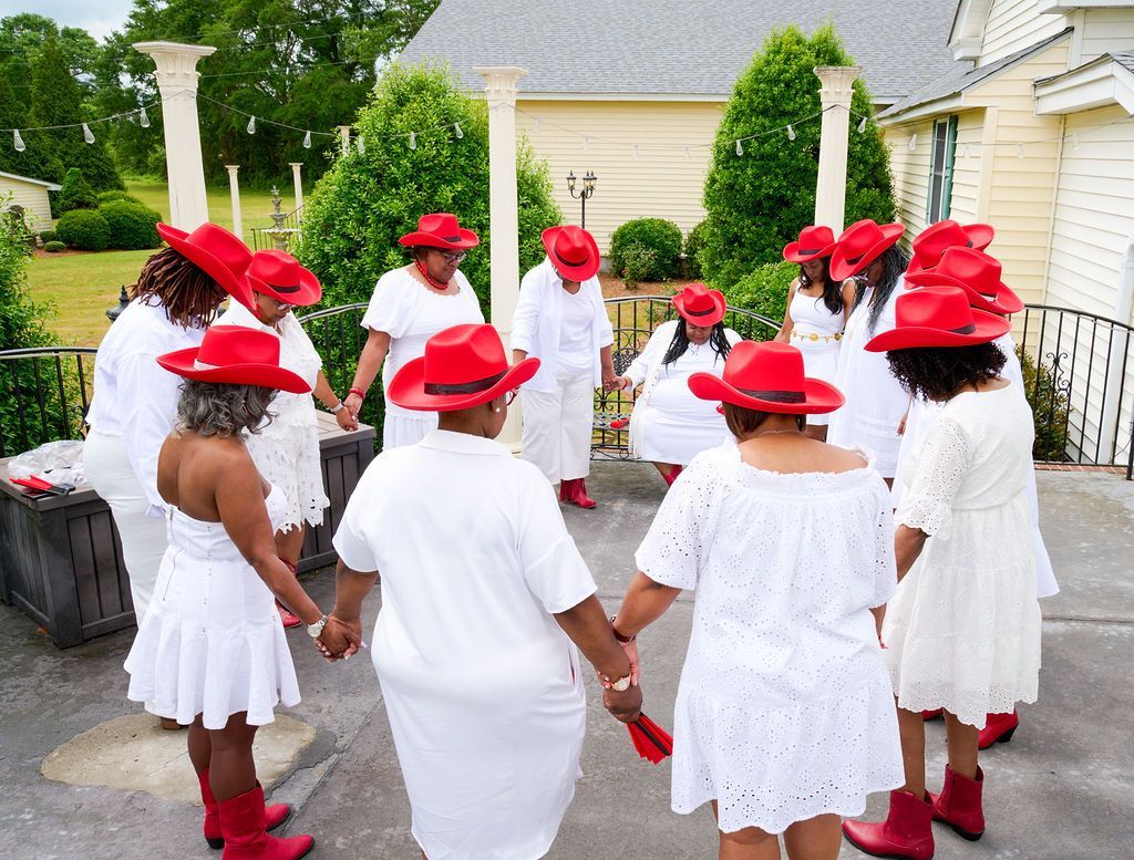 Group of women in white outfits and red hats and boots, holding hands in a circle outdoors.
