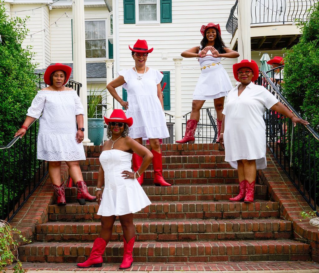 Five people in white dresses and red cowboy hats and boots pose on brick steps.