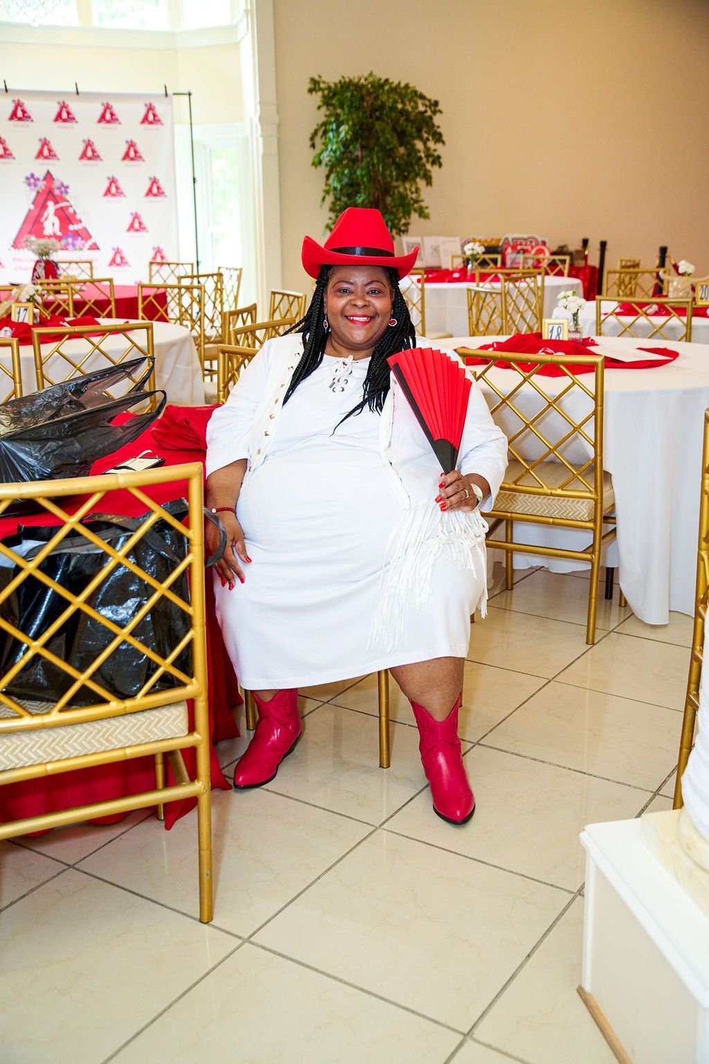 Woman in red cowboy hat and boots, white dress, seated, holding a fan at a decorated table.