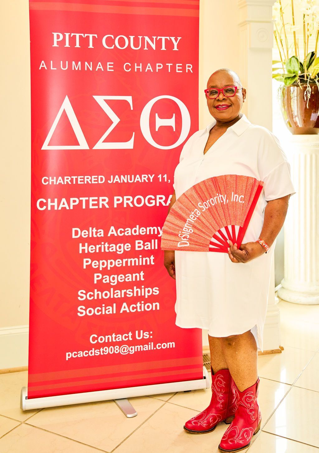 Woman in white dress and red boots poses next to a red banner for Pitt County Alumnae Chapter of Delta Sigma Theta.