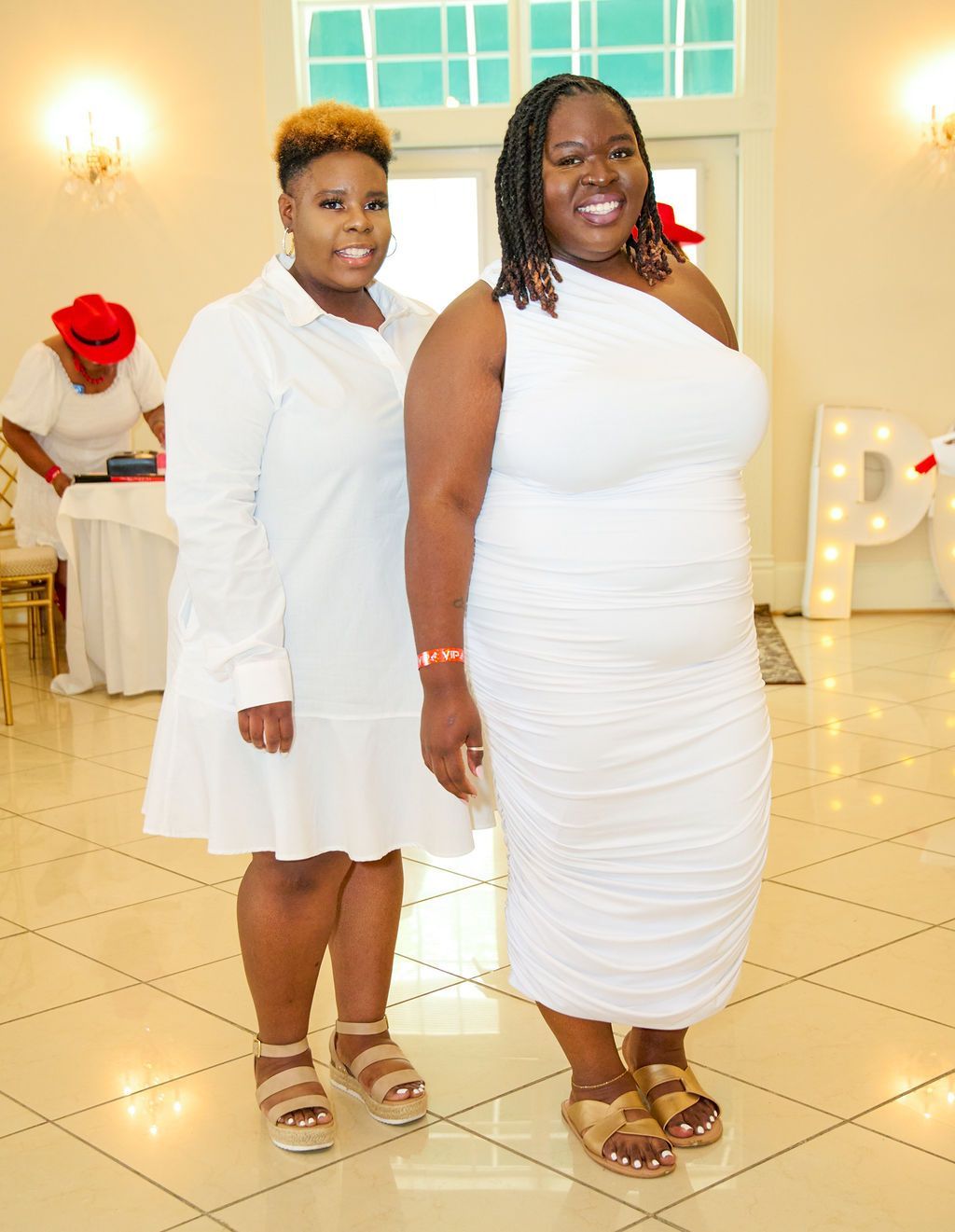 Two women in white dresses smiling at an event; indoor setting with lit-up letters in background.