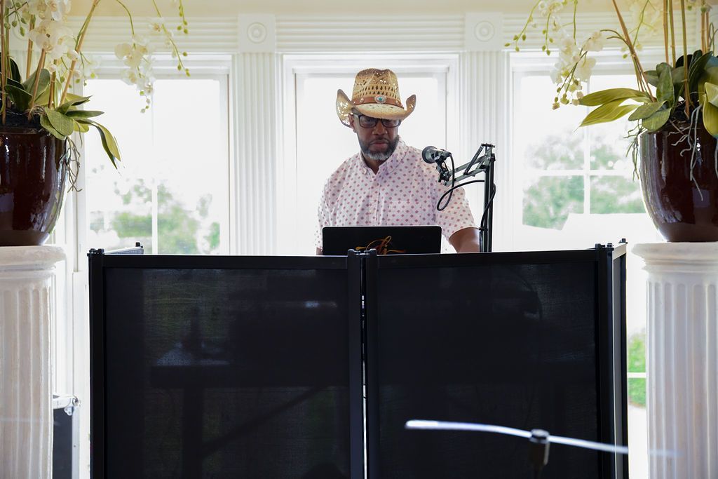 DJ wearing a cowboy hat at a soundboard; windows and floral arrangements in background.