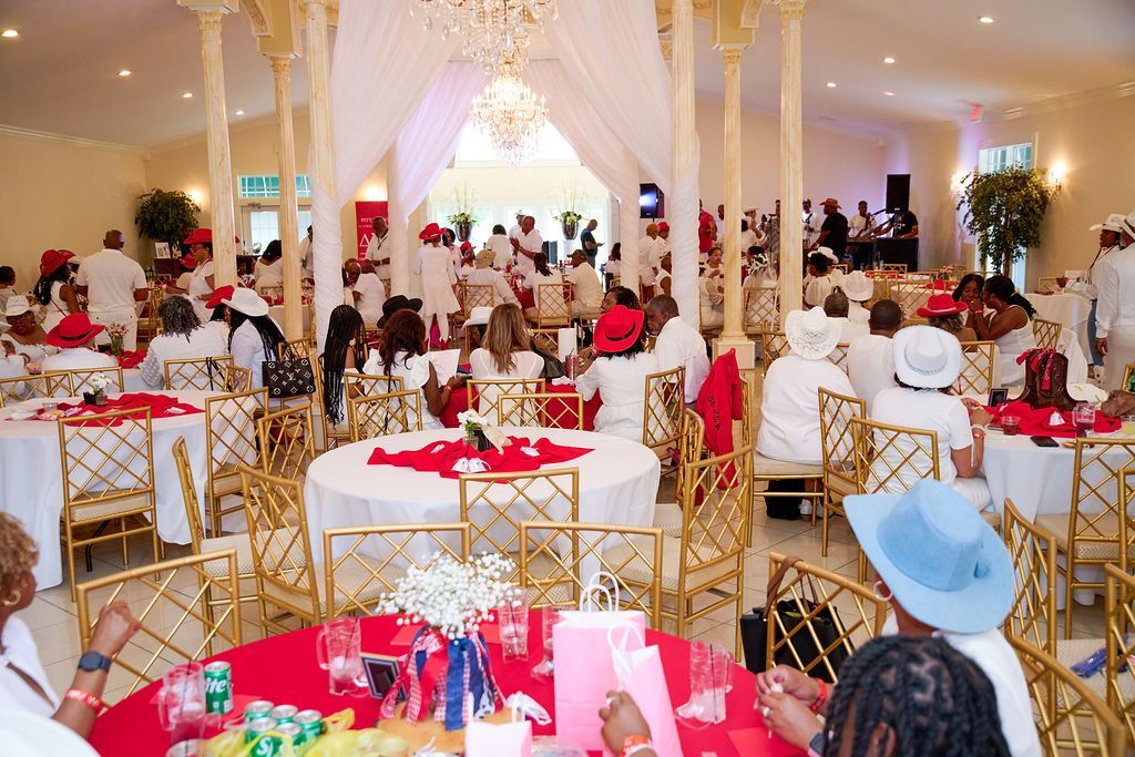 Elegant hall decorated for an event with red and white accents; guests in white attire, dining and socializing.
