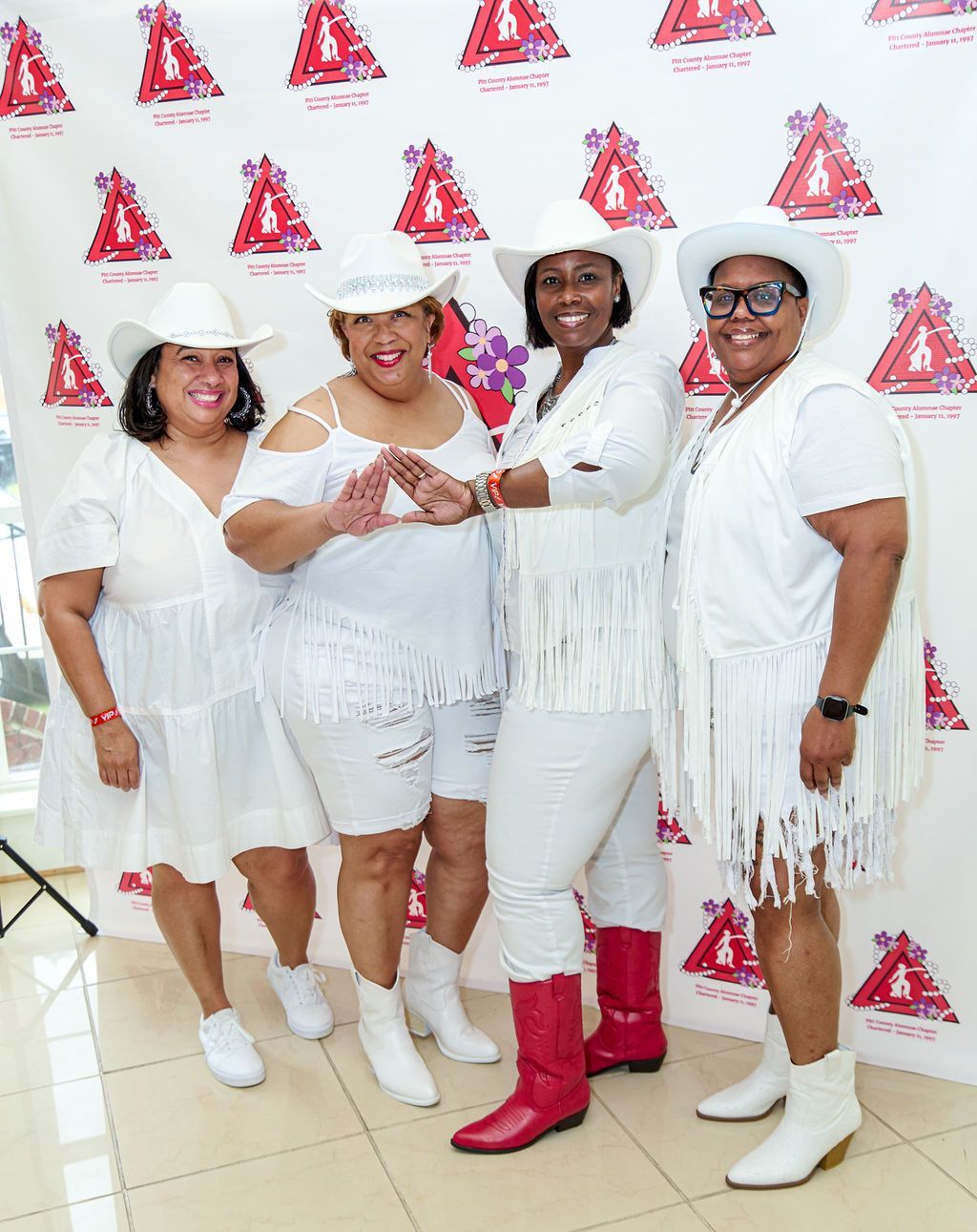 Four people in white outfits and cowboy hats pose in front of a banner. One wears red boots.