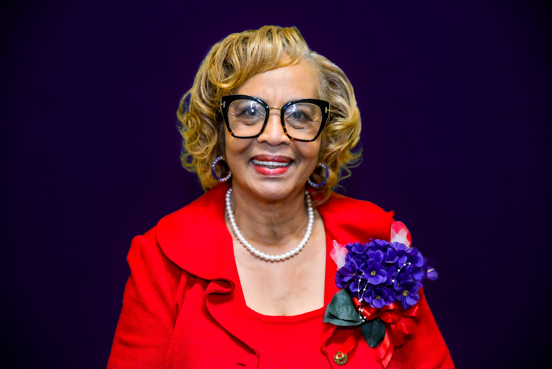 Woman in red suit, wearing glasses and a pearl necklace, smiling against a purple background.