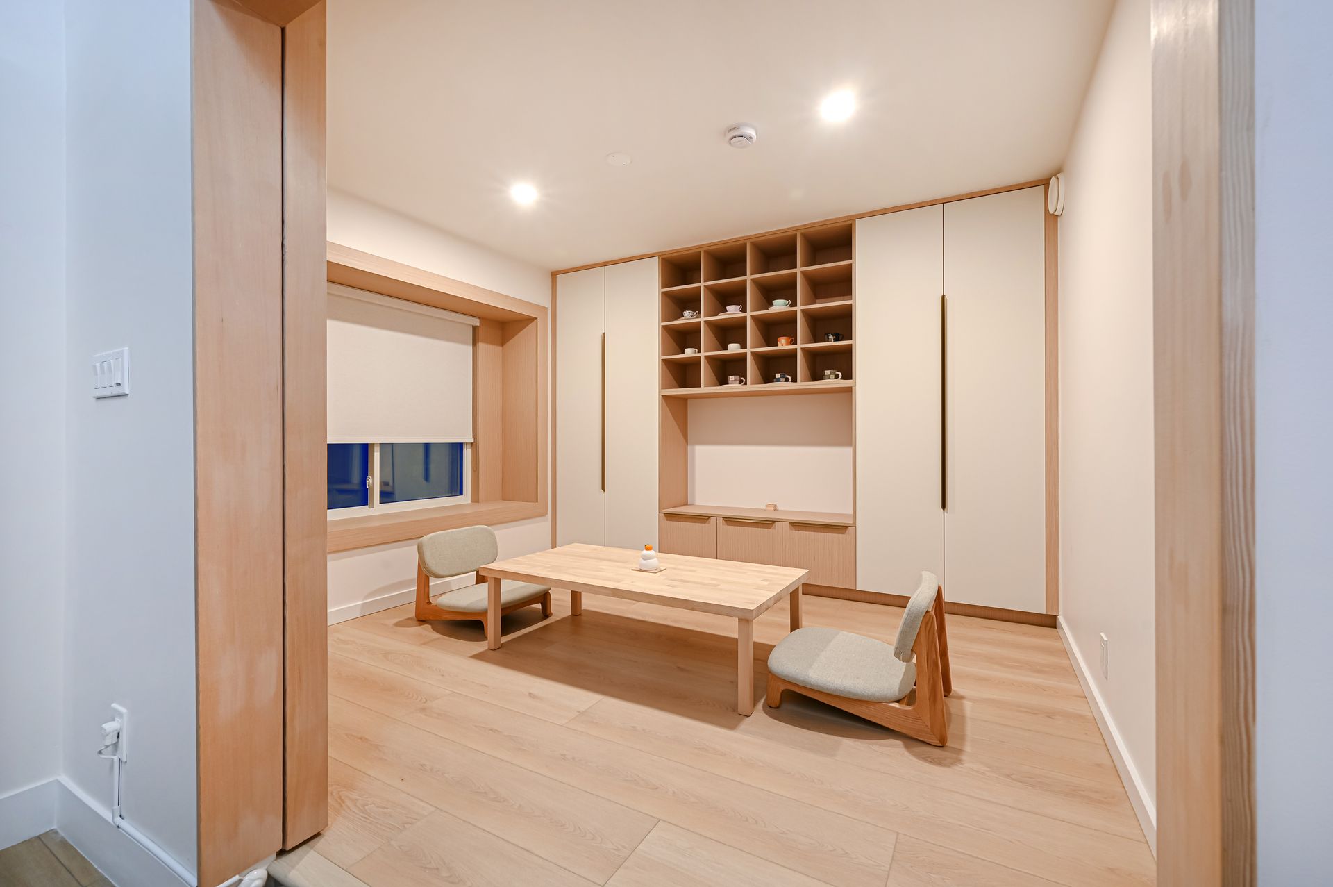 Cozy, light-wood furnished room with a small table, chairs, built-in shelves, and a window.