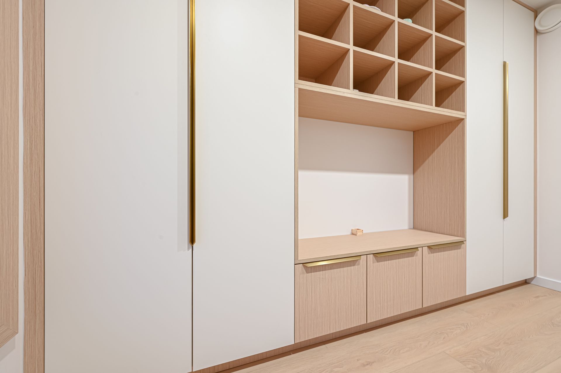 Built-in white and light wood closet with open shelving, drawers, and gold handles against light wood flooring.