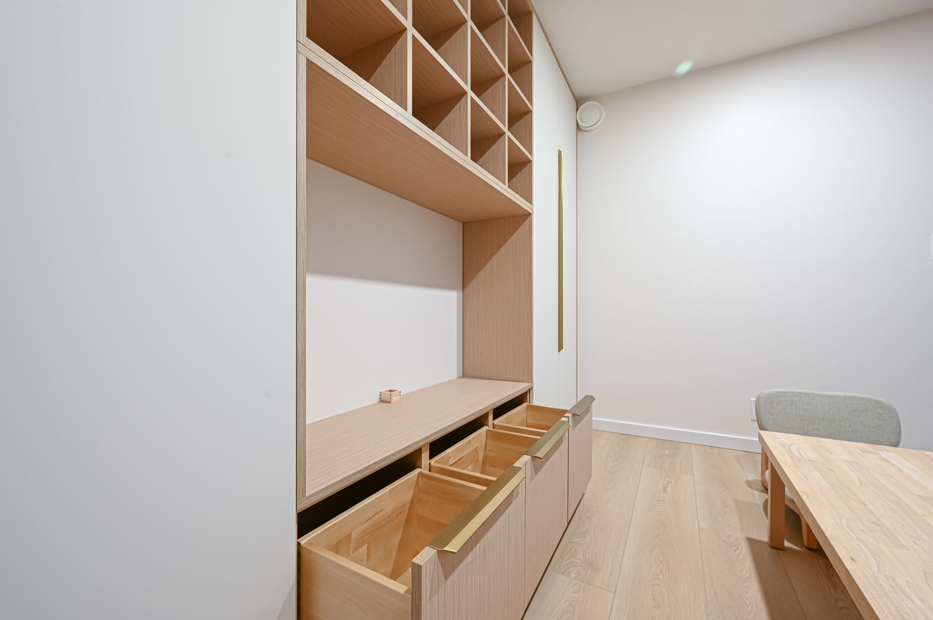 Built-in wooden storage unit with cubbies and drawers in a bright, minimalist room; wooden table and chair.