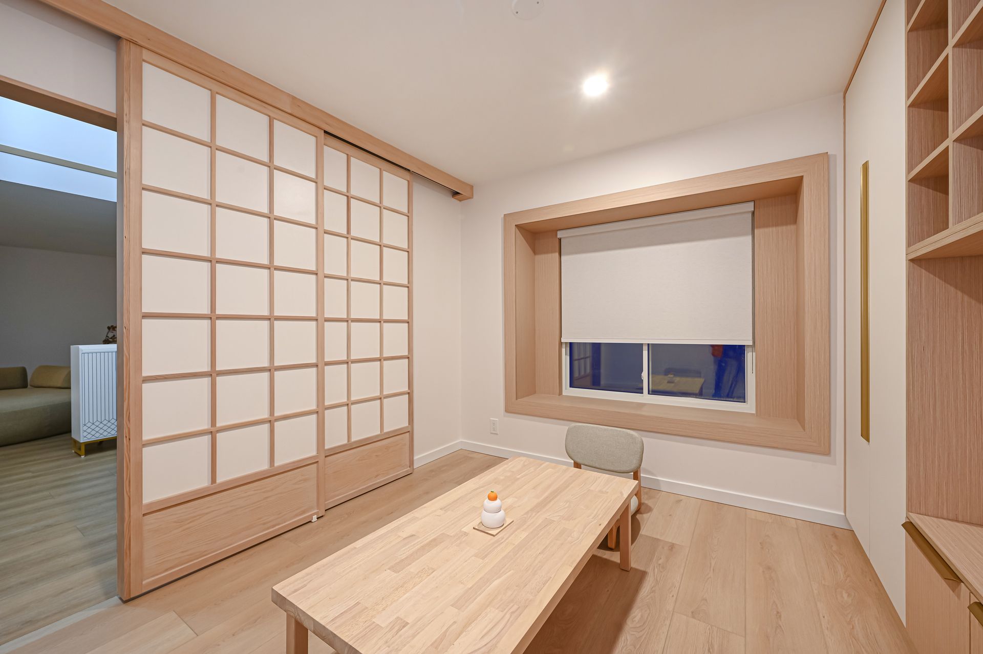 A light-wood furnished room with a sliding paper door, table, window with a closed blind, and shelving.