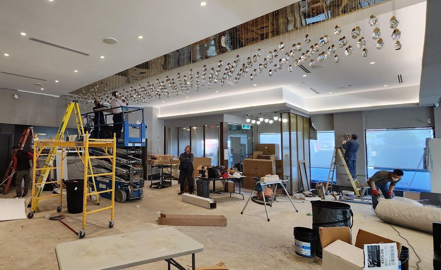 Interior space under construction, featuring workers, scaffolding, and materials. Ceiling lights and mirrored panel are visible.