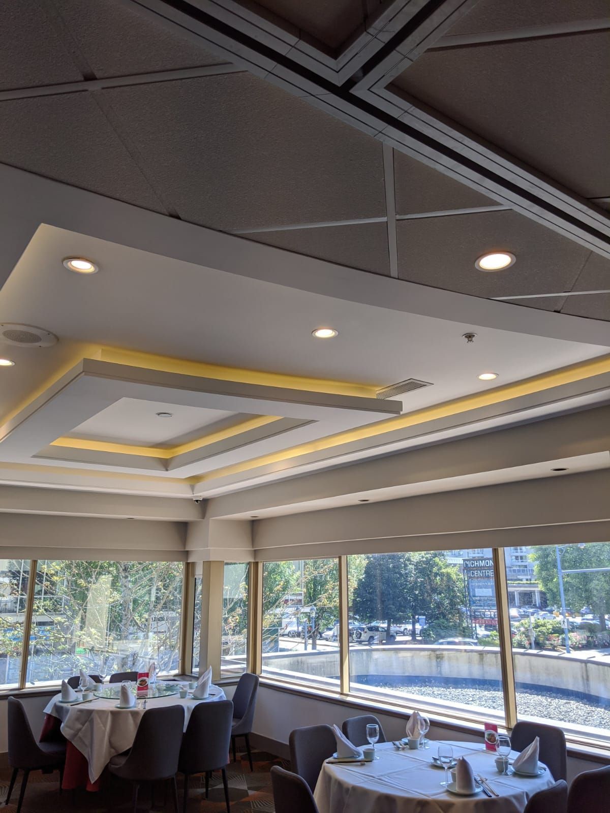 Restaurant interior with round tables set with white tablecloths near large windows, decorative ceiling lights.