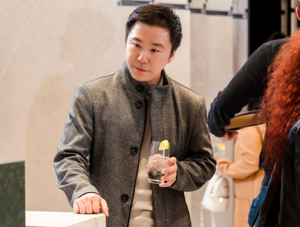 Man in gray jacket holding a drink, looking to the side, at an event.