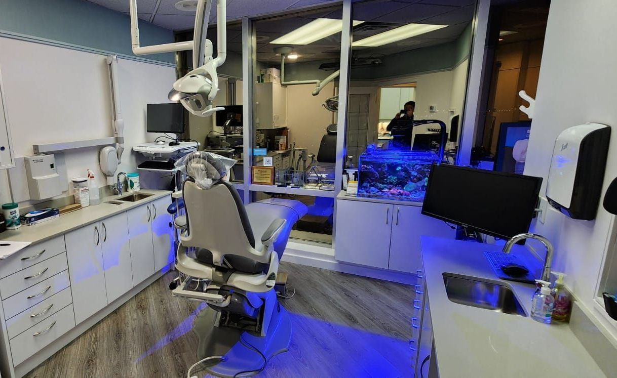 Dental operatory with chair, equipment, and cabinets, seen through a glass partition. Blue light illuminates the floor.