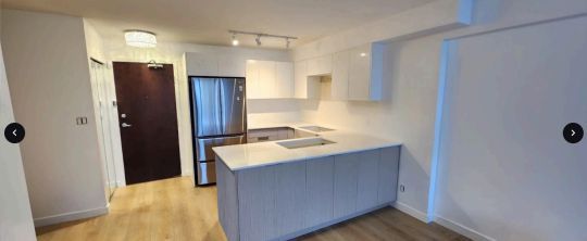 Kitchen with white cabinets, stainless steel fridge, and an island. Brown door. Wooden floor.
