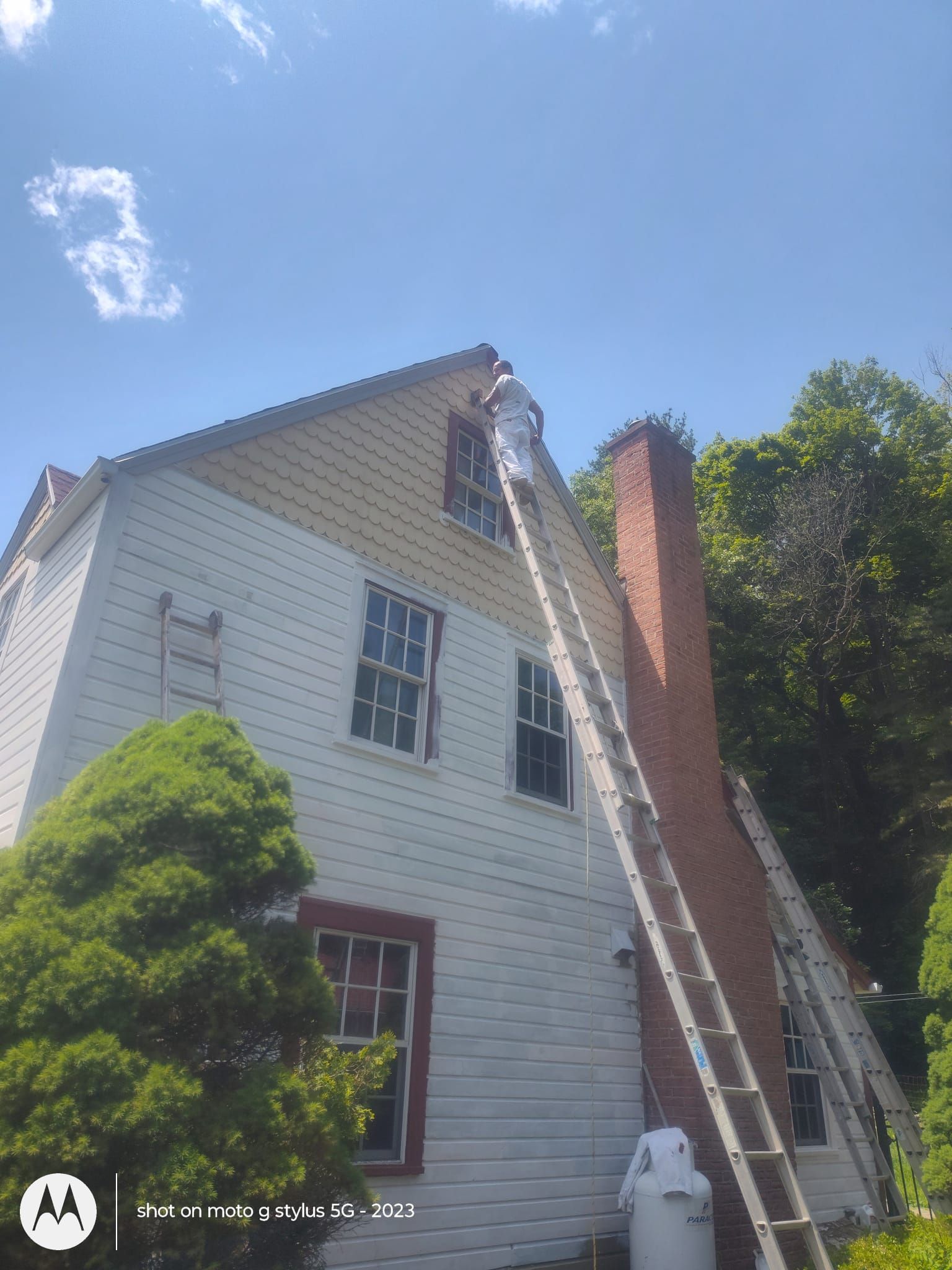 Two-story white house with a brick chimney, ladder leaning against roof, under a blue sky