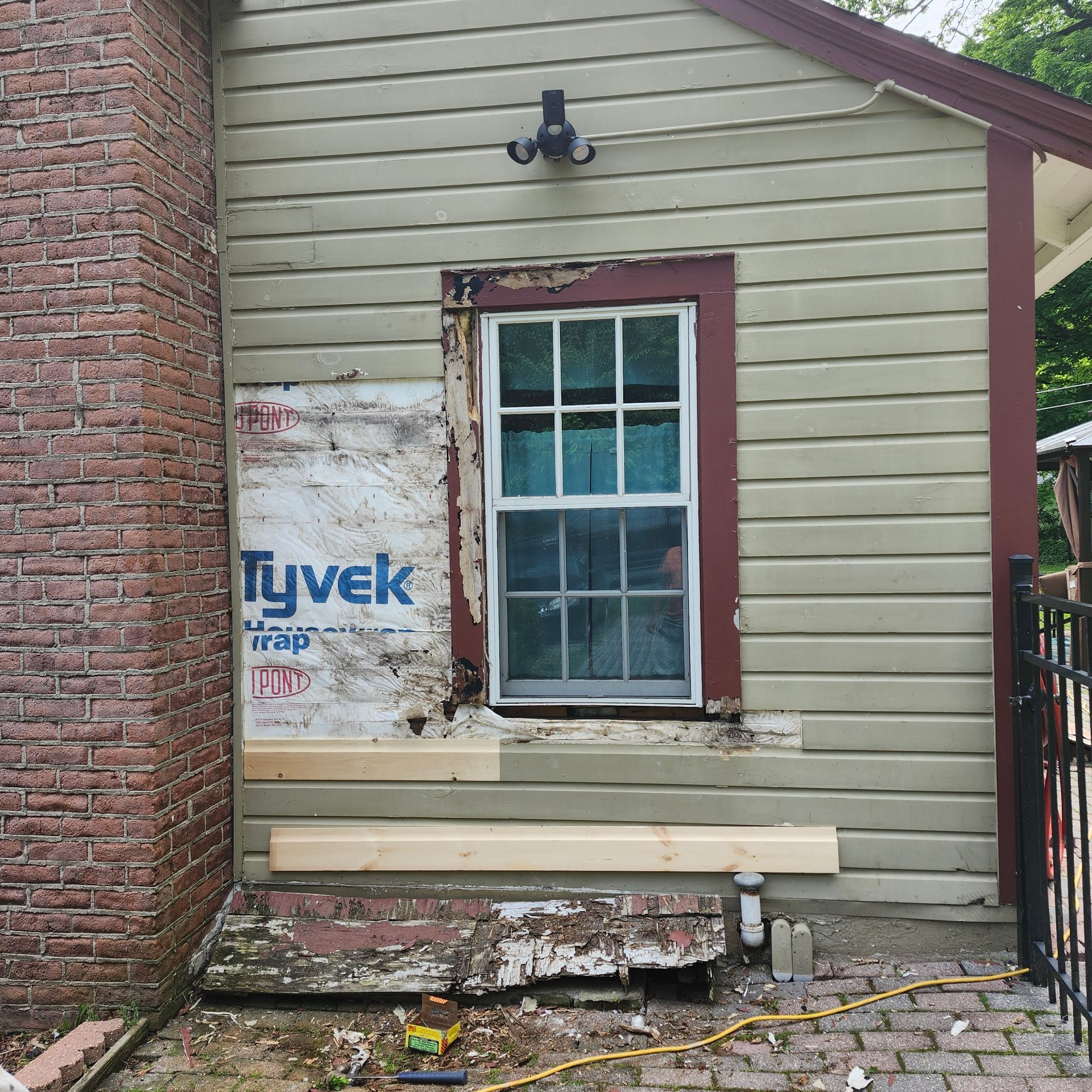 Weathered house exterior with peeling siding, window, brick wall, and construction materials below.