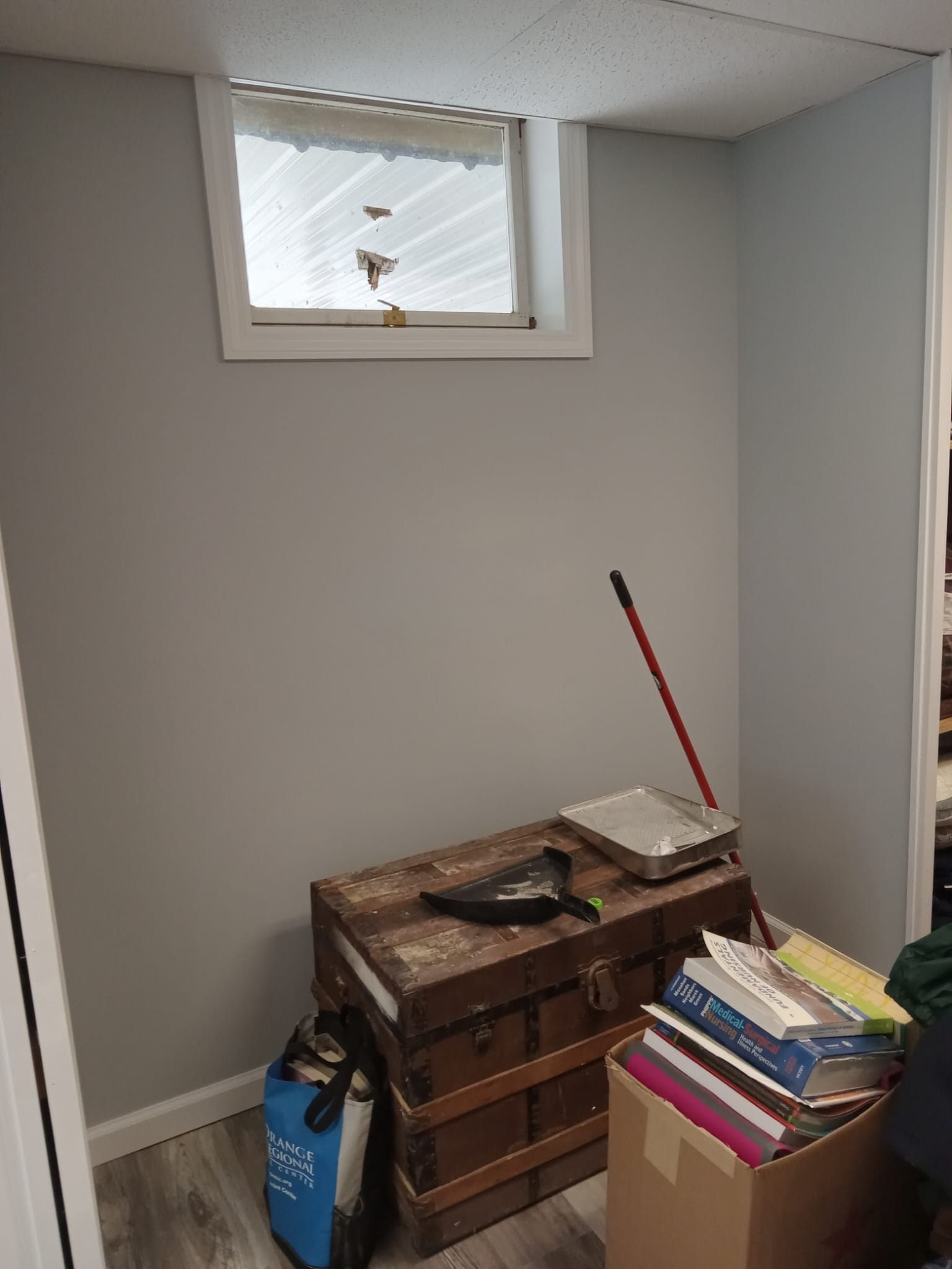 Small room with a window, stacked books, a wooden chest, and a blue bag on the floor.