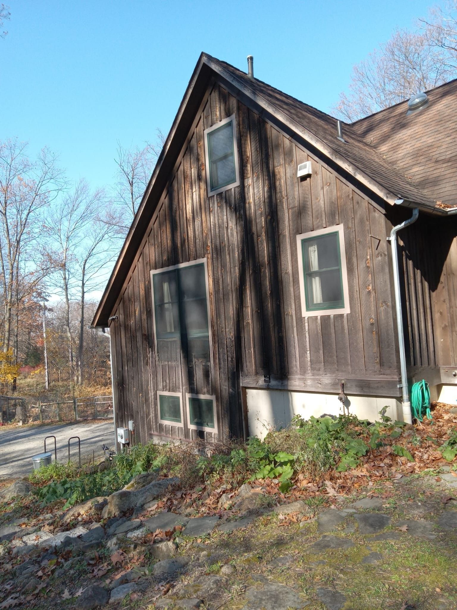 Weathered brown wooden house with steep roof and white trim, seen from the side in a wooded yard.